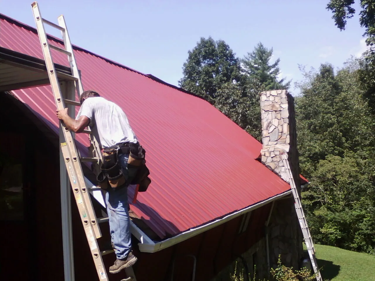Roofer on a ladder beside a red metal roof and stone chimney — Soffit & Fascia Repair by Roofing Brothers in Pineview